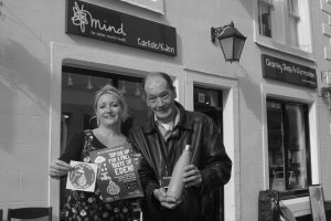 A man and woman outside a Mind charity shop, holding up a Refill sticker and poster, and a reusable water bottle.
