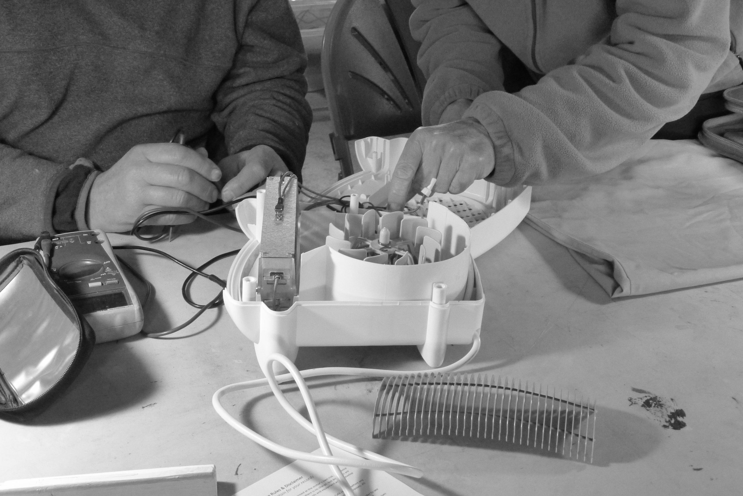 Close up of an electrical item taken apart on a table with the hands of two people