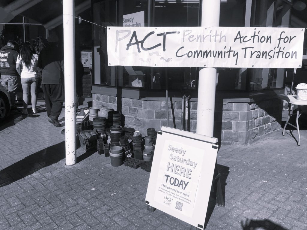 Two signs reading 'Penrith Action for Community Transition' and 'Seedy Saturday Here Today' are displayed outside of a building with flowerpots stacked outside.